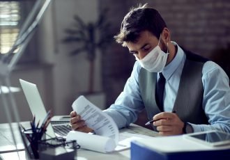 Male entrepreneur analyzing business reports while wearing face mask and working in the office during virus epidemic.